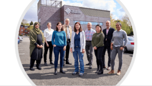 Group photo of nine diverse individuals standing in front of a modern brick building labeled "The BioHub Birmingham" against a partially cloudy sky. The people are dressed in casual attire, with a range of ages, genders, and ethnicities represented. They are standing on a parking lot, with cars visible in the background. The photo is enclosed within a subtle white oval frame.