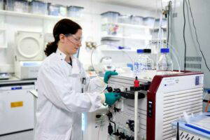 A woman in a lab coat, safety glasses, and teal gloves adjusts dials on a scientific instrument with clear bottles and tubing attached, in a bright laboratory setting.