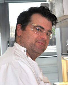 A man with short dark hair and a short beard, wearing clear safety glasses and a white lab coat, smiles slightly while looking towards the viewer. The background includes a window and lab equipment.